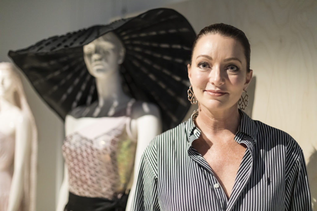 Smiling woman with her hair pulled back in a pony-tail standing in front of a mannequin dressed in a large brimmed hat and sequinned bodysuit; the mannequin is out of focus.